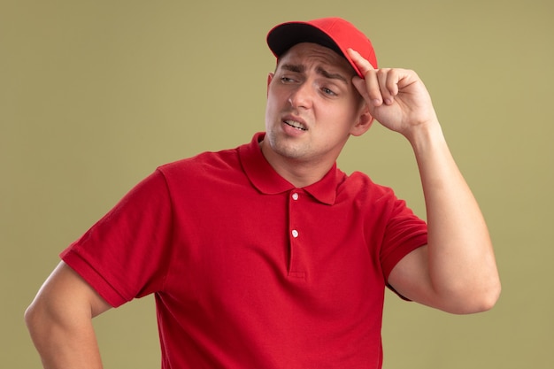 Kostenloses Foto müder blick auf den jungen lieferboten der seite, der uniform und kappenhaltekappe lokalisiert auf olivgrüner wand trägt