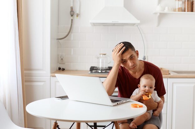 Müder, erschöpfter Mann, gutaussehender Freiberufler, der ein burgunderfarbenes R-Shirt trägt, in der weißen Küche posiert, mit dem Baby in den Händen vor dem Laptop sitzt und Kopfschmerzen hat.