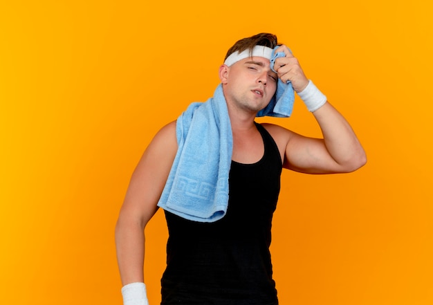 Müder junger hübscher sportlicher Mann, der Stirnband und Armbänder mit Handtuch um den Hals trägt, wischt seinen Schweiß mit Handtuch ab, das auf orange Hintergrund lokalisiert wird