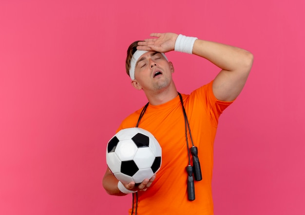Müder junger hübscher sportlicher Mann, der Stirnband und Armbänder mit Springseil um den Hals hält Fußball hält Hand auf Stirn mit geschlossenen Augen lokalisiert auf rosa Hintergrund mit Kopienraum