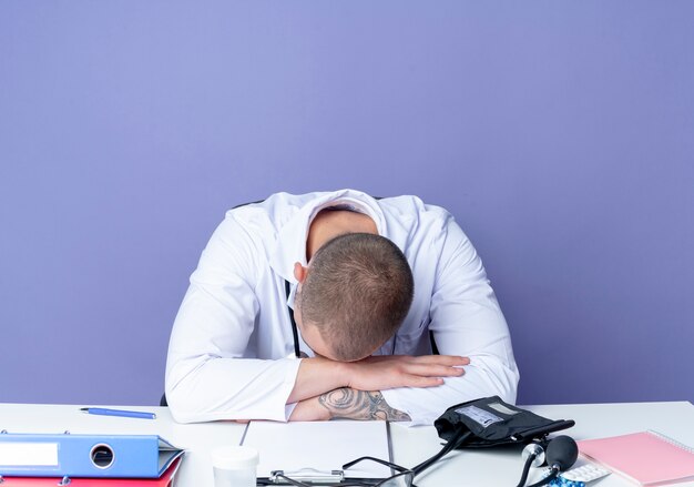 Müder junger männlicher Arzt, der medizinische Robe und Stethoskop trägt, sitzt am Schreibtisch mit Arbeitswerkzeugen, die Hände auf Schreibtisch setzen und Kopf auf Hände lokalisiert auf lila Hintergrund