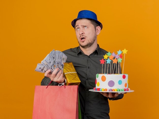 Müder junger party-typ, der blauen hut trägt, der geschenke mit kuchen und geld auf orange isoliert hält