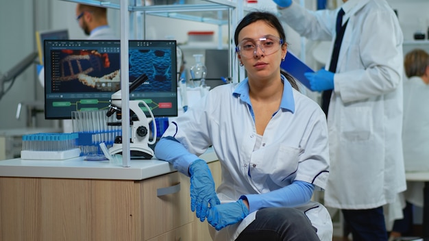 Müder laborarzt, der die kamera im modern ausgestatteten labor lächelt. multiethnisches team, das die virusentwicklung mit hightech- und chemiewerkzeugen für wissenschaftliche forschung und impfstoffentwicklung untersucht.