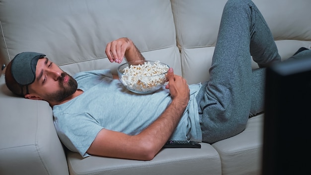 Müder mann, der sich auf dem sofa vor dem fernseher entspannt und popcorn isst, während er eine filmshow sieht