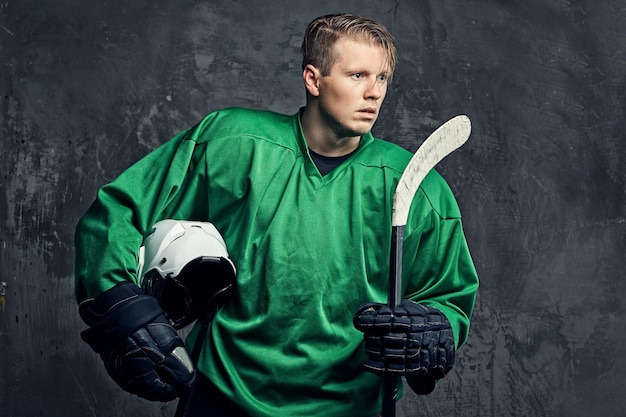 Müder professioneller Hockeyspieler in grüner Sportbekleidung hält einen Hockeyschläger und einen Schutzhelm. Isoliert auf grauem Hintergrund.