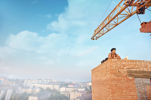 Müder und gutaussehender Baumeister mit Hut, der auf der Mauer auf der Höhe liegt und ruht. Wegschauen. Blauer Himmel mit Wolken in der Sommersaison im Hintergrund. Milch und Brot in der Nähe.