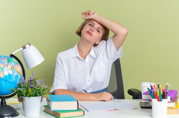 Müdes junges blondes Studentenmädchen, das am Schreibtisch mit Schulwerkzeugen sitzt und in die Kamera schaut und den Arm auf der Stirn hält, isoliert auf olivgrüner Wand
