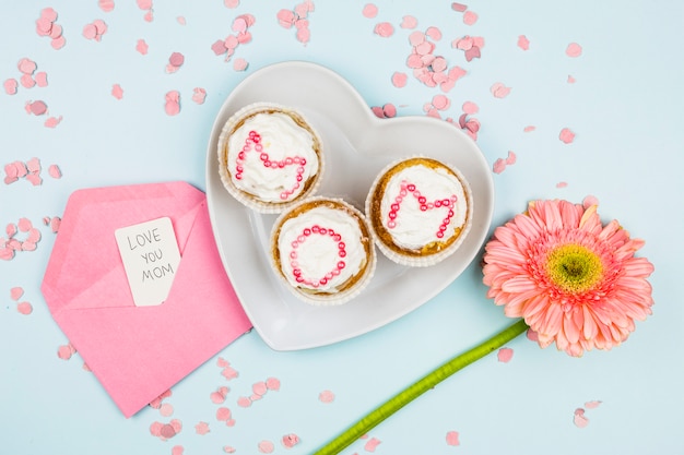 Muffins mit Mutterwort auf Platte nahe Blume und Umschlag mit Tag zwischen Konfetti