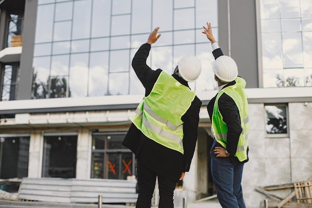 Multi-Rassenbauer stehen im Freien, Rückansicht. Uniform tragen, über Glasneubau sprechen. Arbeiten am Projekt. Stadtinfrastruktur