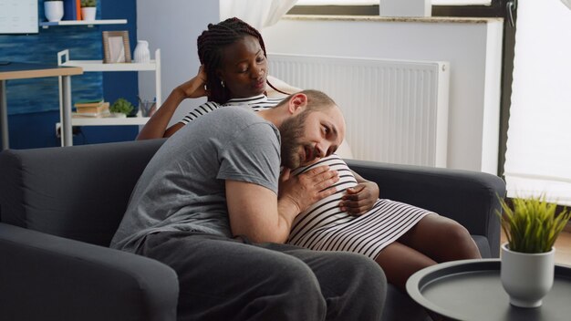 Multiethnische Eltern sprechen über Baby und Elternschaft, während sie zusammen auf der Couch sitzen. Kaukasischer Vater des Kindes, der den Bauch einer afroamerikanischen Frau zu Hause berührt. Interracial Paar