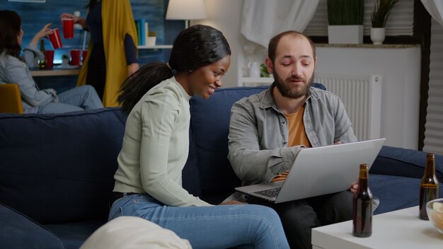 Multiethnische Freunde treffen sich, während sie lustige Online-Videos auf dem Laptop ansehen, der auf dem Sofa im Rücken ruht ...