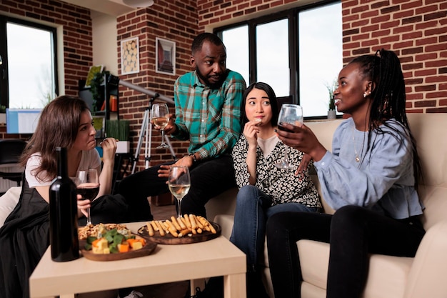 Multiethnische Gruppe von Freunden, die im Wohnzimmer sitzen und gemeinsam eine Weinparty genießen. Junge Leute bei Geburtstagsveranstaltungen trinken alkoholische Getränke, während sie Erinnerungen erzählen.