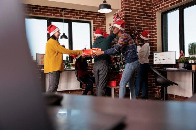 Multiethnische Gruppe von Menschen, die bei der Weihnachtsfeier im Geschäftsbüro Weihnachtsgeschenke austauschen. Kollegen geben am Arbeitsplatz Geschenke, die mit festlichen Weihnachtsornamenten und Lichtern geschmückt sind.