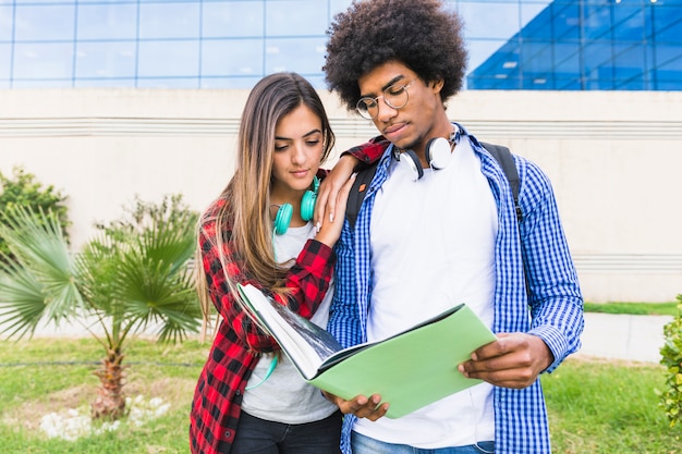 Multiethnische junge paare, die zusammen das buch stehen gegen universitätsgebäude lesen