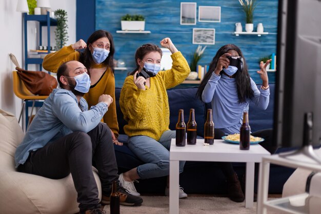 Multiethnische Menschen feiern den Sieg des Videospiels im Wohnzimmer mit Joystick, der eine Gesichtsmaske trägt und in Zeiten des Corona-Ausbruchs soziale Distanz hält. Diverse Freunde, die Bier und Pommes genießen.
