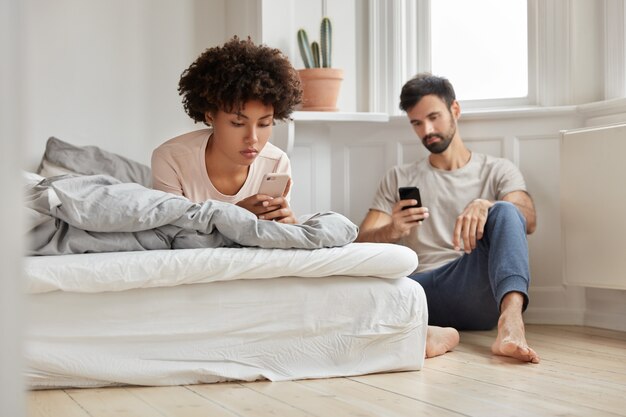 Multiethnische Paare schauen sich Videos über soziale Netzwerke an, halten moderne Mobiltelefone, Frau liegt im Bett, Ehemann sitzt in der Nähe des Bodens, schreibt eine SMS, posiert in einem geräumigen hellen Raum zu Hause. Onlinekommunikation