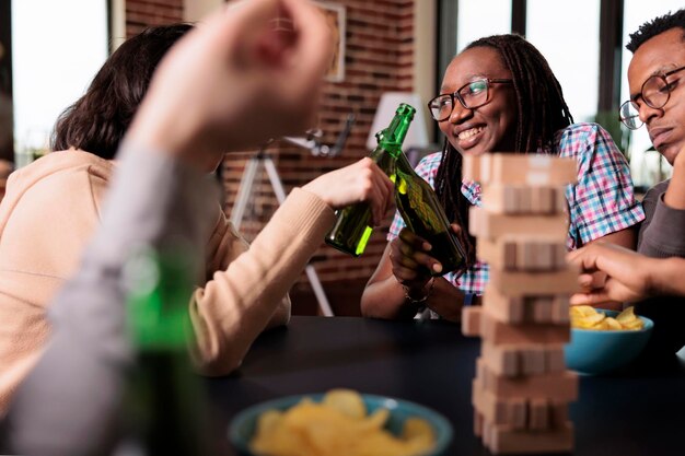 Multikulturelle glückliche Freunde, die gemeinsam alkoholische Getränke trinken, während sie Gesellschaftsspiele spielen. Multiethnische fröhliche Menschen sitzen zu Hause im Wohnzimmer und entspannen sich bei Freizeitaktivitäten.