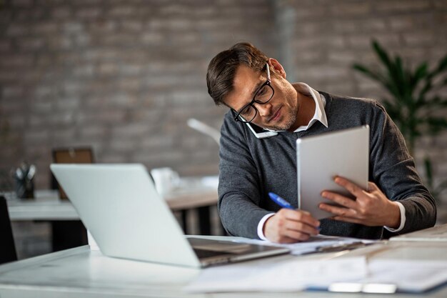 Multitasking-Geschäftsmann, der im Büro arbeitet Er spricht am Telefon, während er sich Notizen macht und ein digitales Tablet verwendet