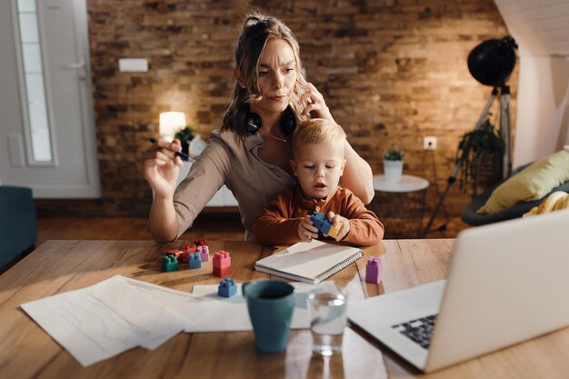 Multitasking-Mutter babysittet ihren kleinen Sohn, während sie von zu Hause aus arbeitet