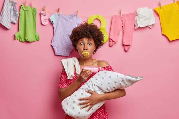 Multitasking-Mutter füttert Neugeborene mit Milch, saugt an der Brustwarze, hält das Kind in eine Decke gewickelt, kümmert sich um ein kleines Kind, hat einen besorgten Ausdruck schockiert, als sie schlechte Nachrichten hört. Elternschaft, Mutterschaft