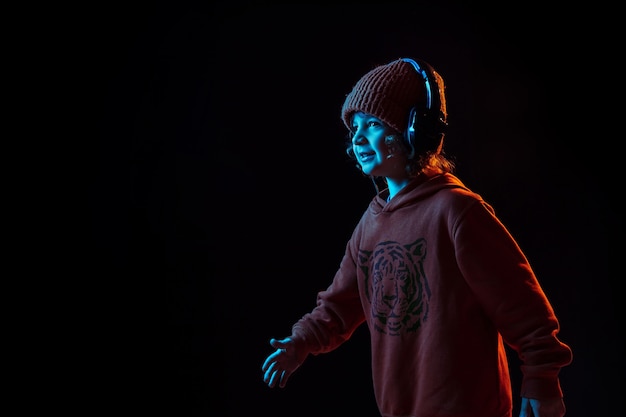 Musik hören und tanzen. Porträt des kaukasischen Jungen auf dunklem Studiohintergrund im Neonlicht. Schönes lockiges Modell. Konzept der menschlichen Emotionen, Gesichtsausdruck, Verkauf, Werbung, moderne Technologie, Gadgets.