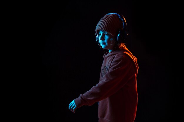 Musik hören und tanzen. Porträt des kaukasischen Jungen auf dunkler Wand im Neonlicht. Schönes lockiges Modell. Konzept der menschlichen Emotionen, Gesichtsausdruck, Verkauf, Werbung, moderne Technologie, Gadgets.