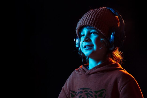 Musik hören und tanzen. Porträt des kaukasischen Jungen auf dunkler Wand im Neonlicht. Schönes lockiges Modell. Konzept der menschlichen Emotionen, Gesichtsausdruck, Verkauf, Werbung, moderne Technologie, Gadgets.