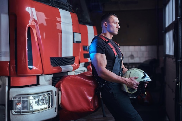 Muskulöser Feuerwehrmann, der einen Schutzhelm in einer Garage einer Feuerwehr hält, sich auf ein Feuerwehrauto stützt und nach draußen schaut