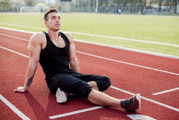 Muskulöser junger männlicher Athlet, der auf dem entspannenden Leichtathletikfeld sitzt