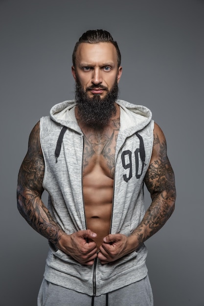 Kostenloses Foto muskulöser tätowierter mann mit bart in grauen zipuo-hoodies. isoliert auf grauem hintergrund.