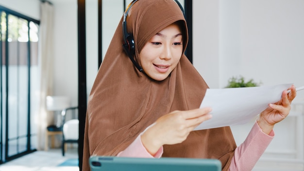 muslimische dame tragen kopfhörer mit digitaler tablette sprechen mit kollegen über verkaufsbericht in konferenz-videoanrufen, während sie von zu hause in der küche arbeiten.