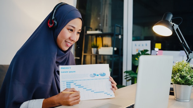 muslimische dame tragen kopfhörer mit laptop sprechen mit kollegen über verkaufsbericht in konferenz-videoanrufen, während sie nachts vom home-Office arbeiten