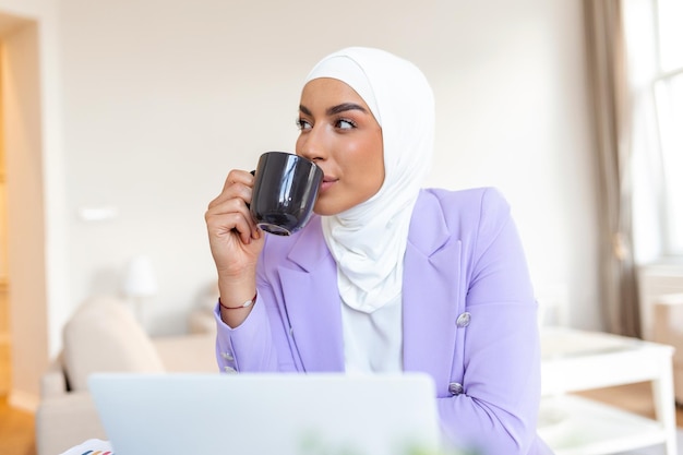 Muslimische Frau, die mit Computer arbeitet Arabische junge Geschäftsfrau, die zu Hause an ihrem Schreibtisch sitzt, an einem Laptop-Computer arbeitet und Kaffee oder Tee trinkt