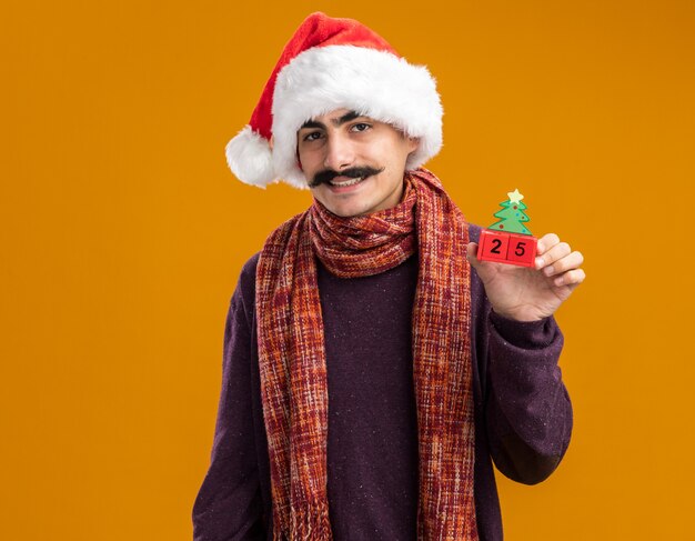 Mustachioed Mann, der Weihnachtsweihnachtsmütze mit warmem Schal um seinen Hals trägt, der Spielzeugwürfel mit Datum fünfundzwanzig betrachtet Kamera mit Lächeln auf Gesicht, das über orange Hintergrund steht