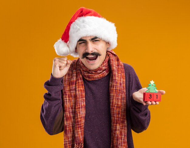 Mustachioed Mann, der Weihnachtsweihnachtsmütze mit warmem Schal um seinen Hals trägt, der Spielzeugwürfel mit Neujahrsdatum hält, das Faust und glückliches und aufgeregtes Stehen über orange Hintergrund ballt