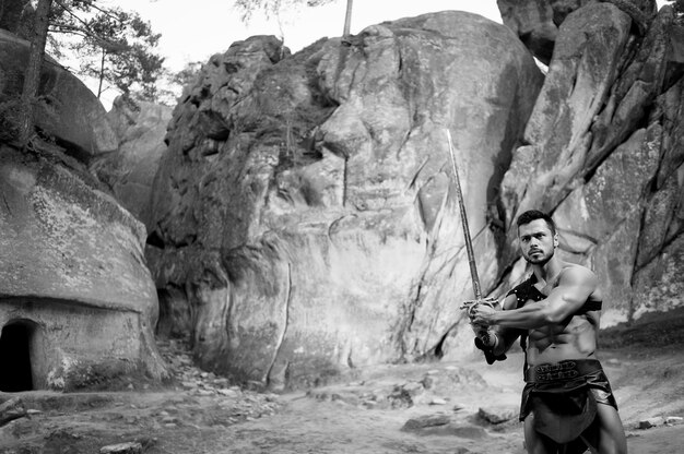 Mutiges Herz. Monochromes Porträt eines jungen Kriegers mit einem atemberaubenden, athletischen, kraftvollen Körper, der bereit ist, mit einem Schwert zu kämpfen, das in der Nähe der Felsen posiert