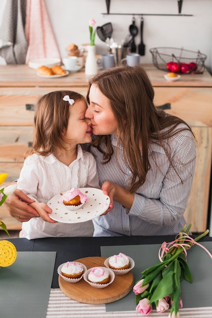 Mutter, die Tochter mit kleinem Kuchen küsst