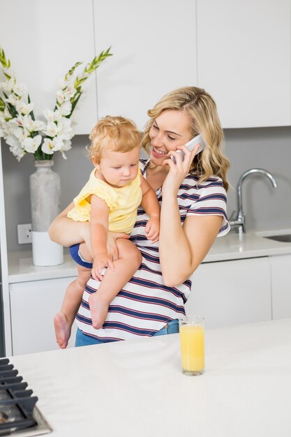 Mutter im Gespräch über Handy, während ihr Baby in der Küche hält