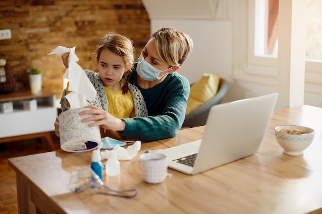 Mutter mit Gesichtsmaske gibt ihrer kleinen Tochter zu Hause eine Schachtel Taschentücher