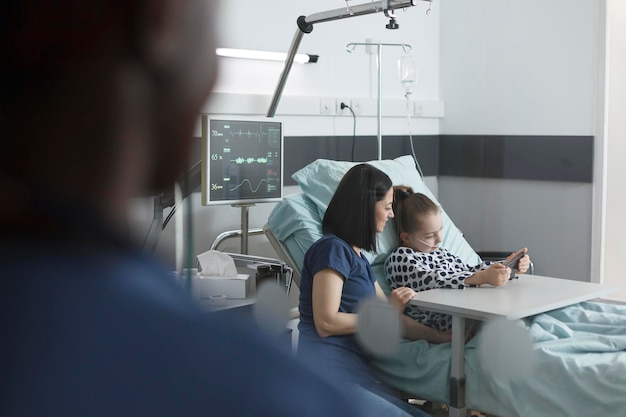 Mutter sitzt neben Patientenbett im Zimmer der Kinderklinik, während süßes kleines Mädchen auf dem Smartphone spielt. Fröhlicher junger Patient, der Handy-Unterhaltung genießt, während er in der Klinik-Aufwachstation sitzt.