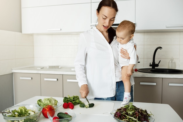 Mutter tragen Baby und bereiten gesundes Essen zu, hacken Gemüse