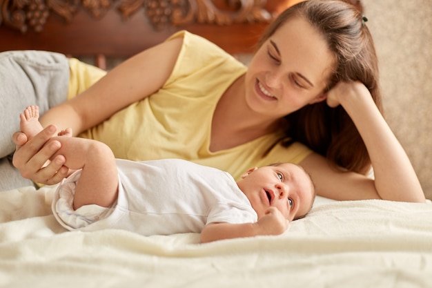 Mutter und Baby, die auf Bett auf weißer Decke liegen, lächelnde Mama, die gelbes T-Shirt trägt, genießen, Zeit mit ihrem neugeborenen Kind zu verbringen, Säugling, der weg schaut, um äußere Dinge zu studieren.