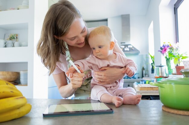 Mutter und kleine Tochter kochen zusammen zu Hause und beobachten Rezepte auf Tablette. Kinderbetreuung oder Kochen zu Hause Konzept