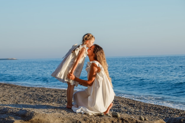 Mutter und Tochter, die tagsüber im weißen Kleid am Meer umarmen.