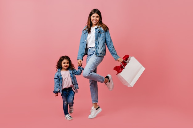 Mutter und Tochter in Jeans Händchen haltend. Studioaufnahme der glücklichen Familie, die auf rosa Hintergrund aufwirft.