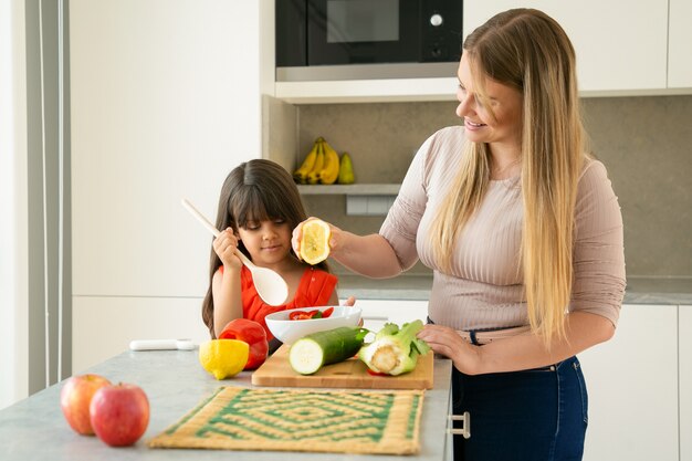 Mutter und Tochter kochen Gemüse zum Abendessen am Küchentisch. Mädchen und ihre Mutter Dressing Salat in Schüssel mit der Hälfte der Zitrone. Familienkochen oder gesundes Ernährungskonzept