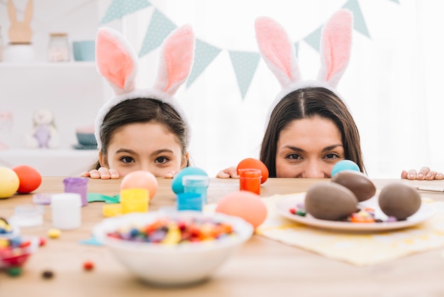 Mutter und Tochter spähen hinter den Tisch mit Ostereiern; Süßwaren und Farben