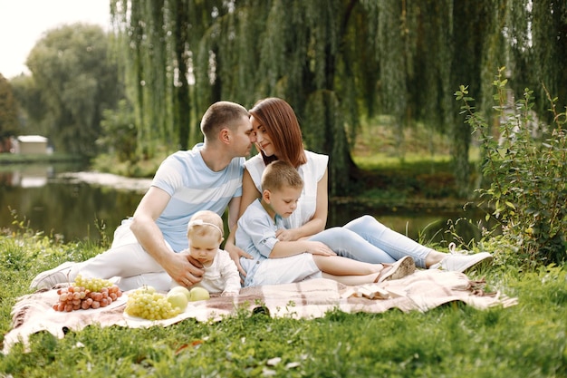 Mutter, Vater, älterer Sohn und kleine Tochter sitzen auf einer Picknickdecke im Park. Familie in weiß-hellblauer Kleidung