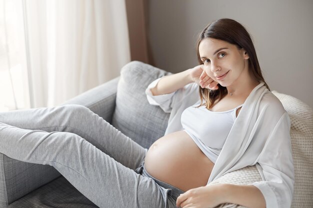 Mutterschaft ist Wunder Porträt einer glücklichen, gutaussehenden jungen zukünftigen Mutter mit langen dunklen Haaren, die auf einem Sessel ruht, den Kopf auf den Arm lehnt und mit süßem Ausdruck in die Kamera lächelt und sich ausruht