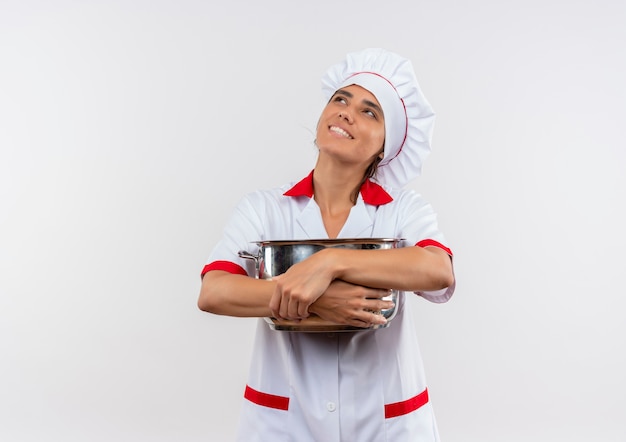 Nach oben schauender lächelnder junger weiblicher Koch, der Kochuniform umarmte Topf auf isolierter weißer Wand mit Kopienraum trägt